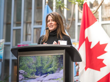 A woman stands behind a podium outside giving a speech with a canadian flag behind her