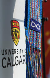 A blue Ceinture Fléchée (Métis arrow sash) featuring the Métis flag hangs in front of the UCalgary logo. In the background a tepee is visible.