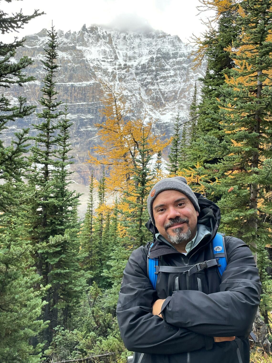 Mauricio Soto-Rubio on a hike, posing in front of coniferous trees and a snowy mountain.